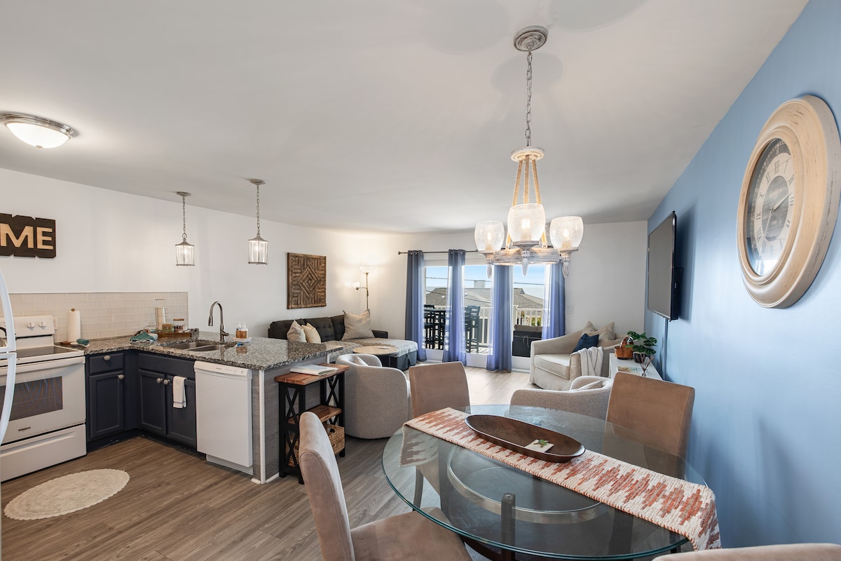 An open-concept living and dining area features a modern kitchen with dark cabinetry and a circular dining table surrounded by cushioned chairs. Natural light fills the space through large sliding glass doors, leading to a covered deck with views of the ocean.