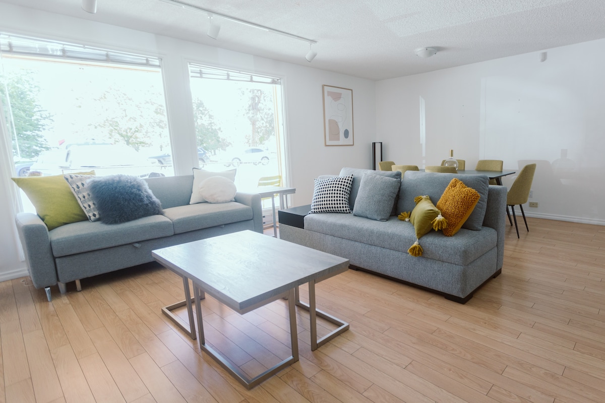 A spacious living area features two modern couches in light grey, one adorned with textured cushions in green and yellow. A minimalist coffee table sits center, while natural light floods in through large windows facing a serene outdoor scene.