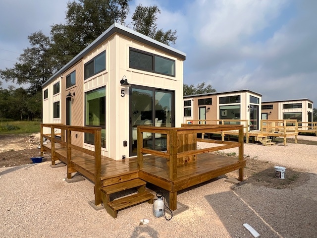 A tiny home is presented with a spacious wooden deck and steps leading to the entrance. Large windows allow for ample natural light, while multiple tiny homes are visible in the background, each designed with a modern exterior.