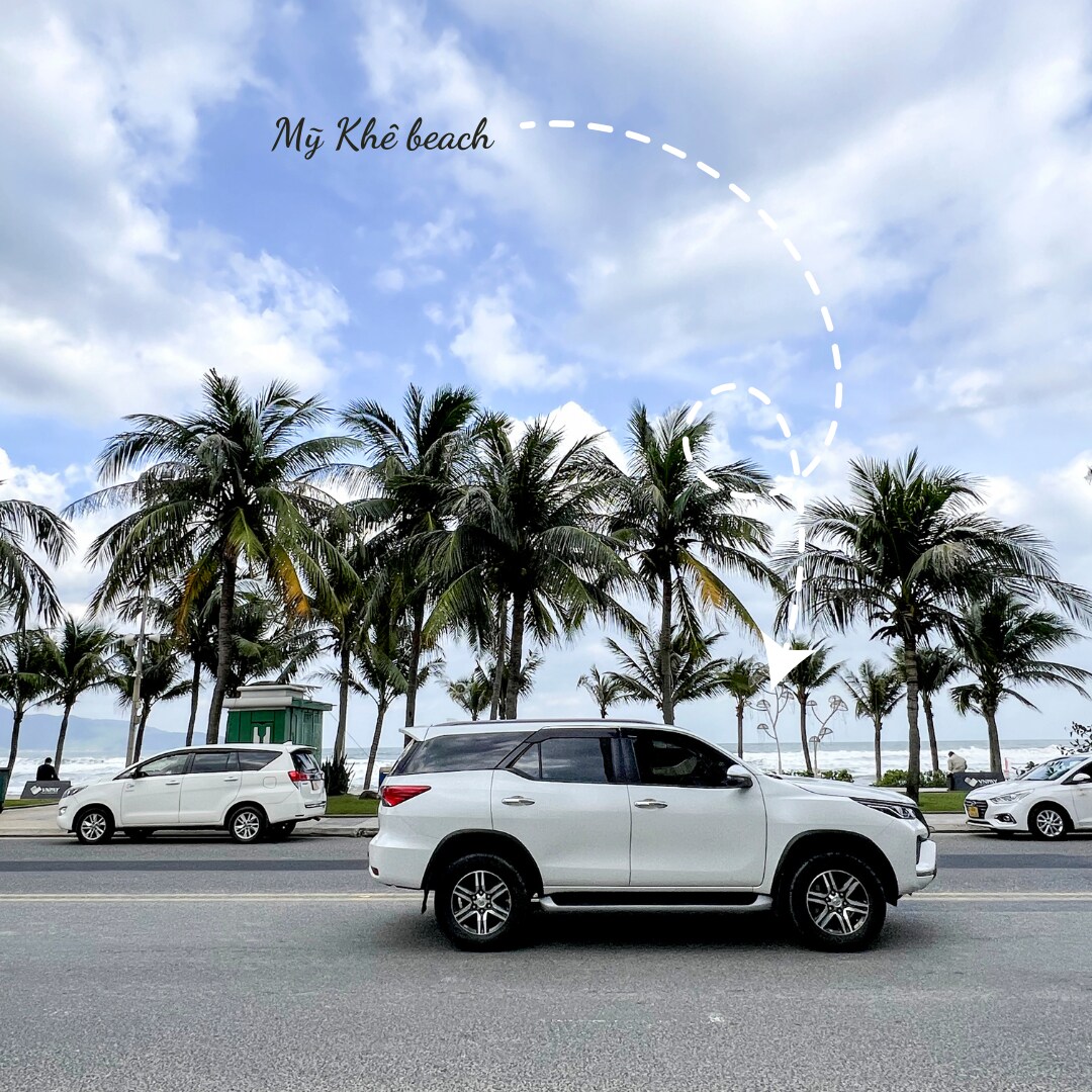 A white SUV is parked along a road lined with palm trees, with the beach in the background. The sky is partly cloudy, and the scene captures a blend of natural and urban elements near Mỹ Khê beach.