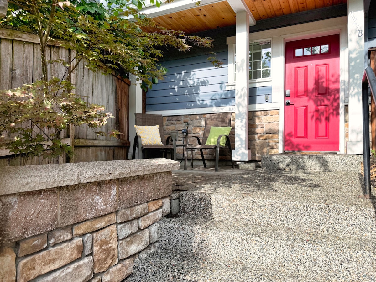 A welcoming entryway is presented, featuring a vibrant red door and two outdoor chairs with cushions. The natural stone steps lead to a covered porch, surrounded by greenery and a wooden fence, creating a cozy atmosphere for relaxation.