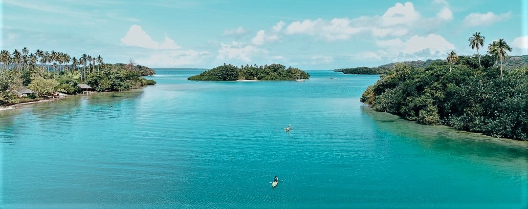 The serene lagoon waters display vibrant turquoise hues, surrounded by lush green islands and palm trees. Kayakers can be seen paddling peacefully across the calm surface, highlighting the area’s natural beauty and tranquil atmosphere.