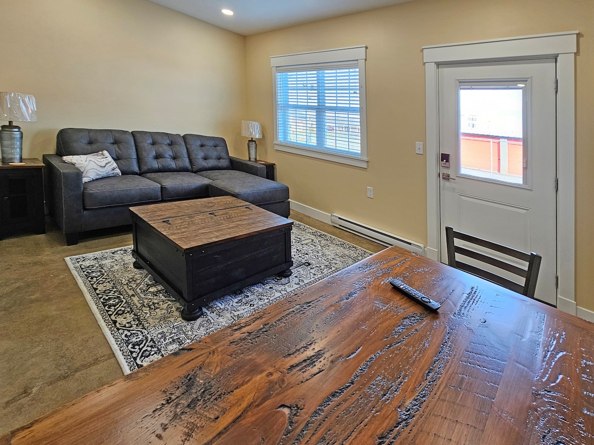 A well-lit living area is displayed, featuring a spacious gray sofa and a wooden coffee table positioned on a patterned rug. A dining table for two is visible in the foreground, while a door with a window provides access to the outside.