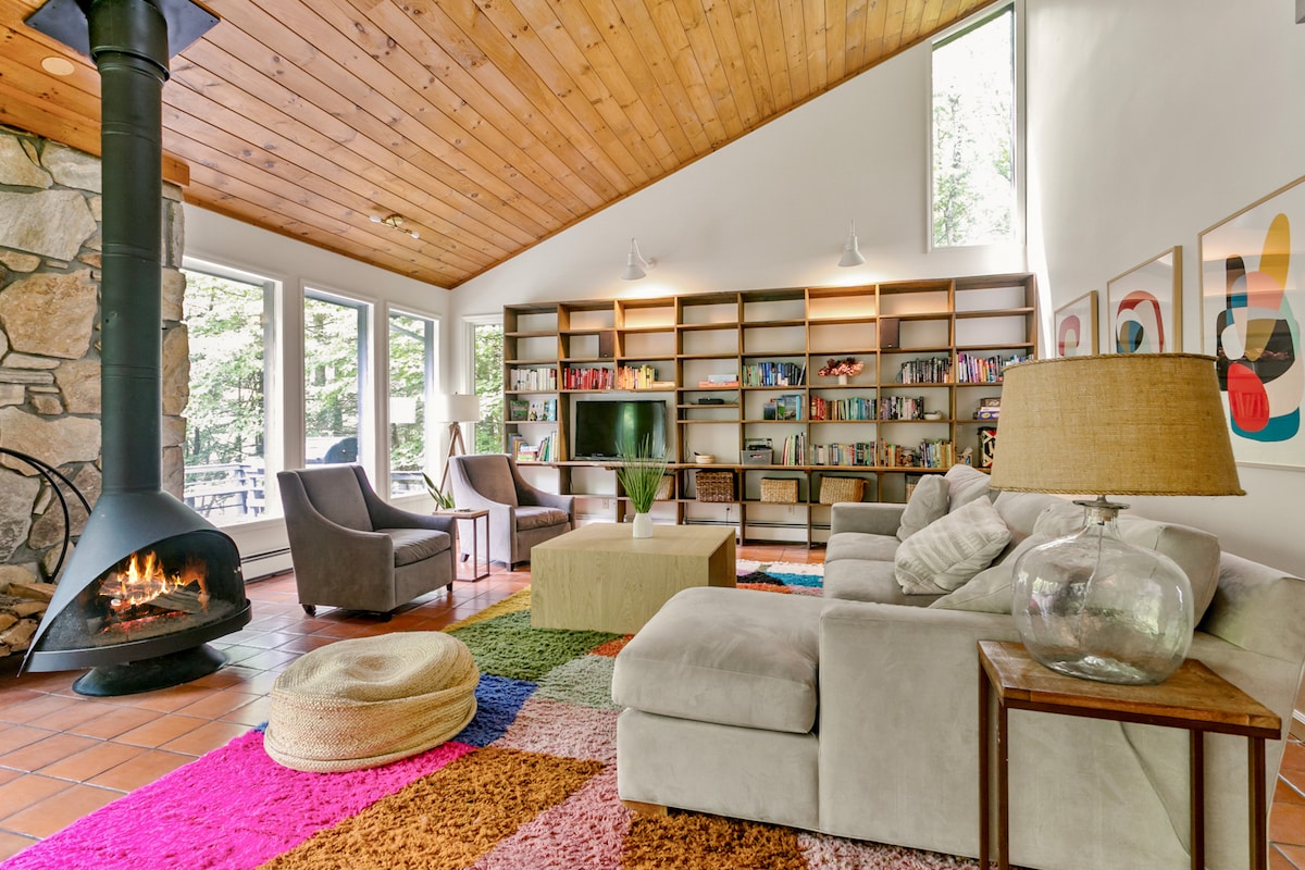 A spacious living room features a stone fireplace, surrounded by bookshelves filled with various books. Large windows allow natural light to brighten the area, which includes a cozy sectional sofa and a colorful area rug. Two armchairs provide additional seating, enhancing the inviting layout.