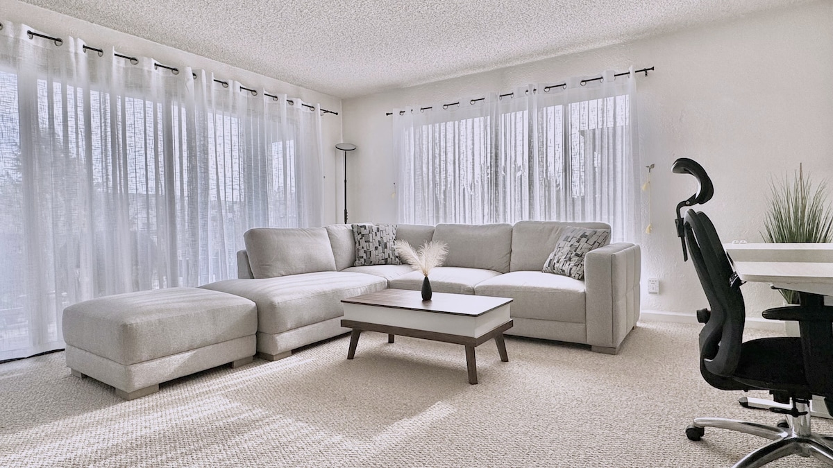 A spacious living area is showcased with a light grey sectional sofa oriented around a low coffee table. Sheer white curtains frame large windows, allowing natural light to fill the room. A desk chair and a small plant are positioned nearby, contributing to a minimalist ambiance.