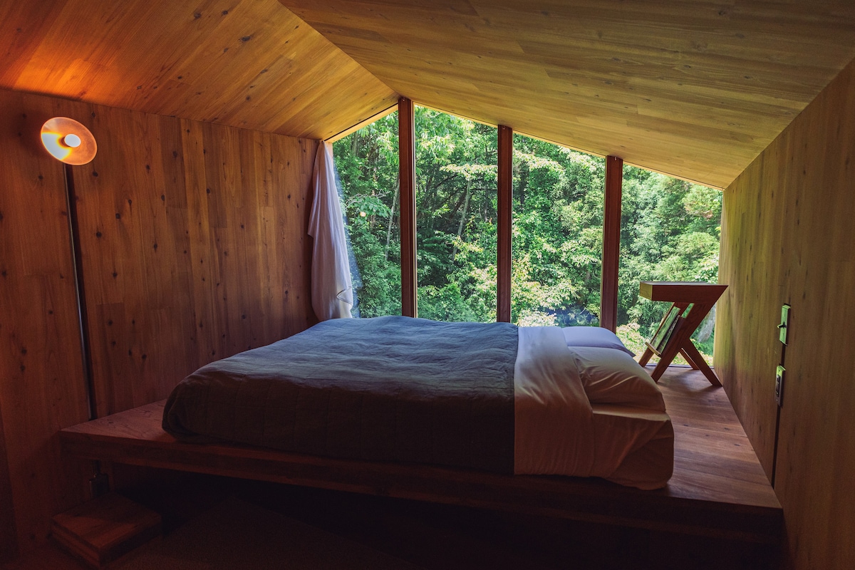 An inviting bedroom features a spacious bed adorned with crisp linens. Large windows overlook lush greenery, allowing natural light to fill the room. A wooden chair is positioned nearby, complementing the natural wood accents of the walls and ceiling.