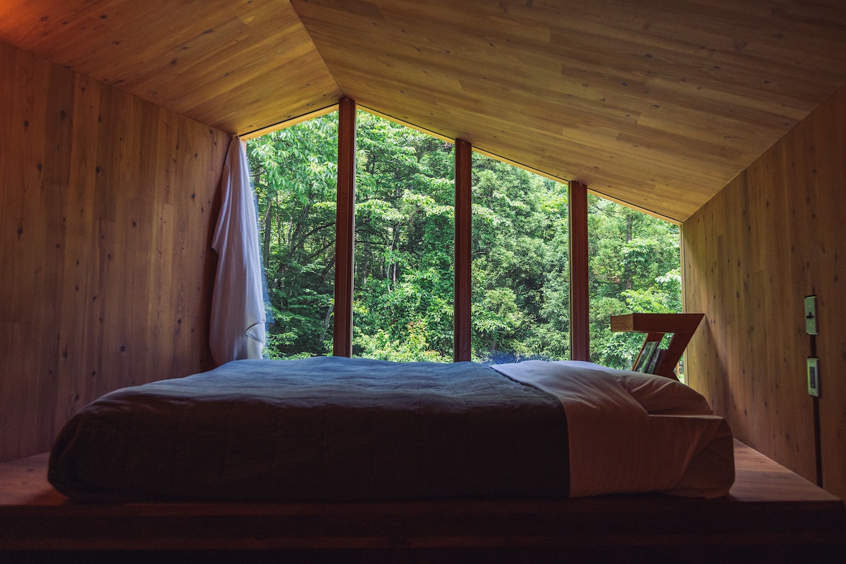 A spacious bed is positioned against a backdrop of large windows, offering views of the surrounding greenery. The wooden walls and ceiling create a natural, warm environment, while soft light illuminates the room, enhancing the connection to the outdoors.