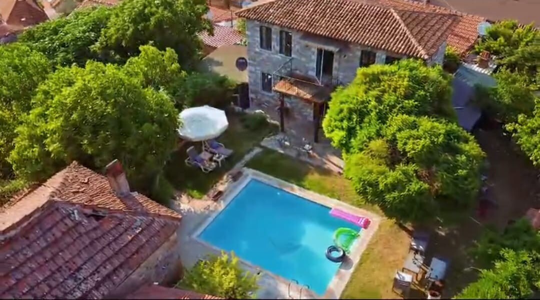 An aerial view features a private stone house surrounded by greenery and trees. A tranquil pool is visible in the spacious backyard, complemented by lounge chairs and an umbrella. The rustic tile rooftops of nearby structures add charm to the serene landscape.
