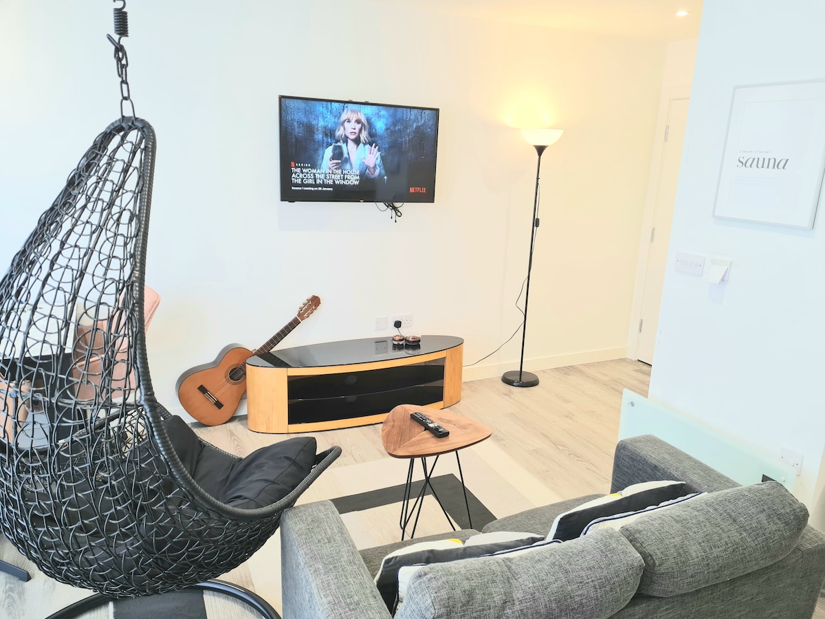 A modern living area features a hanging chair and a sleek sofa, accompanied by a television mounted on the wall. A wooden coffee table holds a remote control. A guitar rests against the wall, with ambient light provided by a standing lamp.