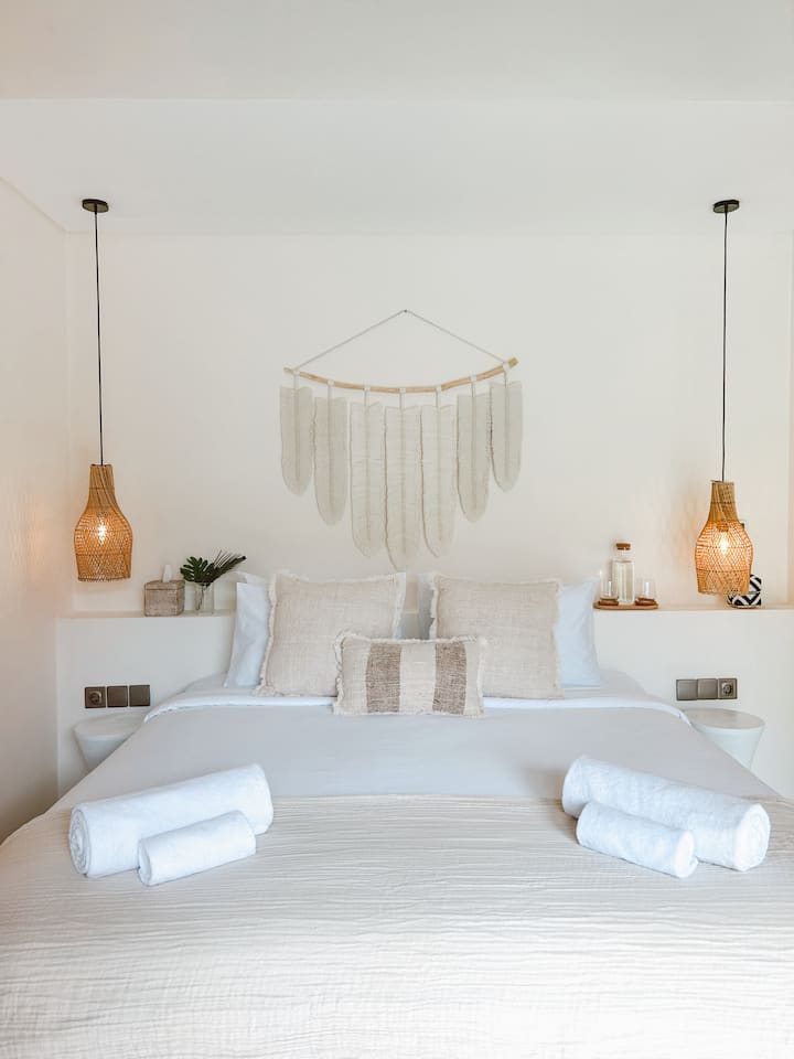 Quarto iluminado e sereno com cama king size, decoração natural, toques de vime, luminárias suspensas e acesso direto a uma área externa exuberante. Mistura perfeita de conforto e elegância tropical.
