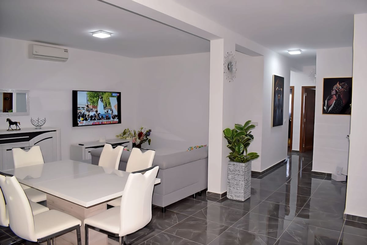 A bright living area is displayed, featuring a modern dining table surrounded by six white chairs. A comfortable gray sofa is positioned nearby, with a flat-screen television mounted on the wall. Decorative plants and framed artwork enhance the welcoming ambiance.
