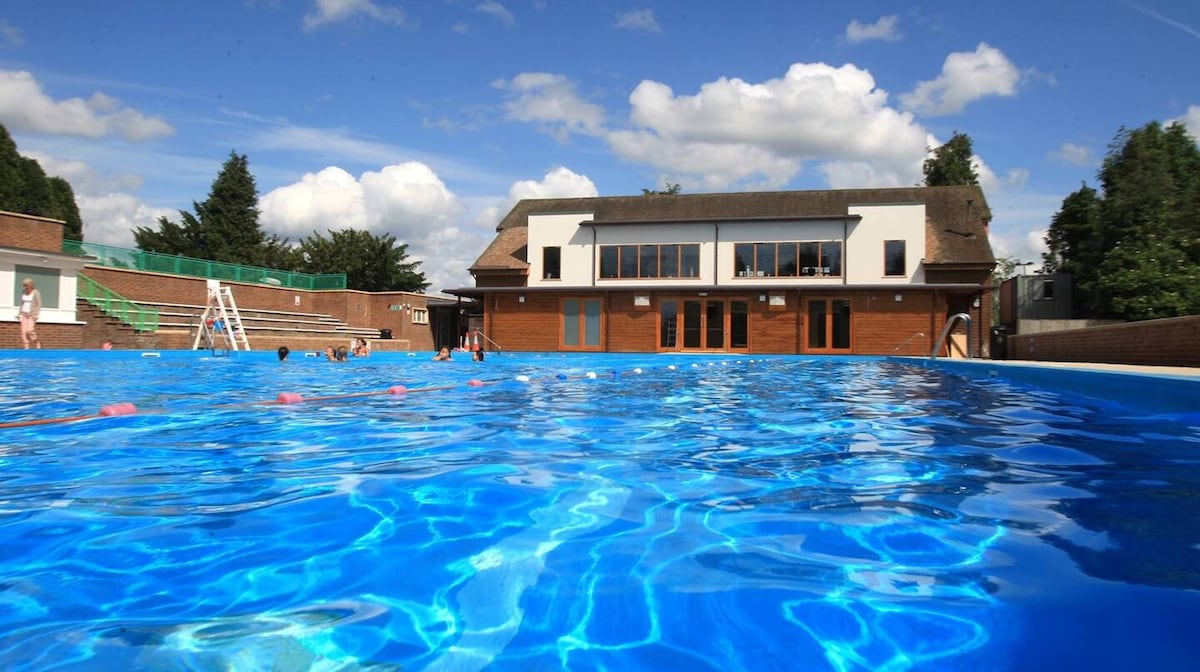 A large outdoor swimming pool is visible in the foreground, characterized by clear, bright blue water. A modern building with large windows sits in the background, surrounded by green trees under a partly cloudy sky. Guests can be seen enjoying the pool area.
