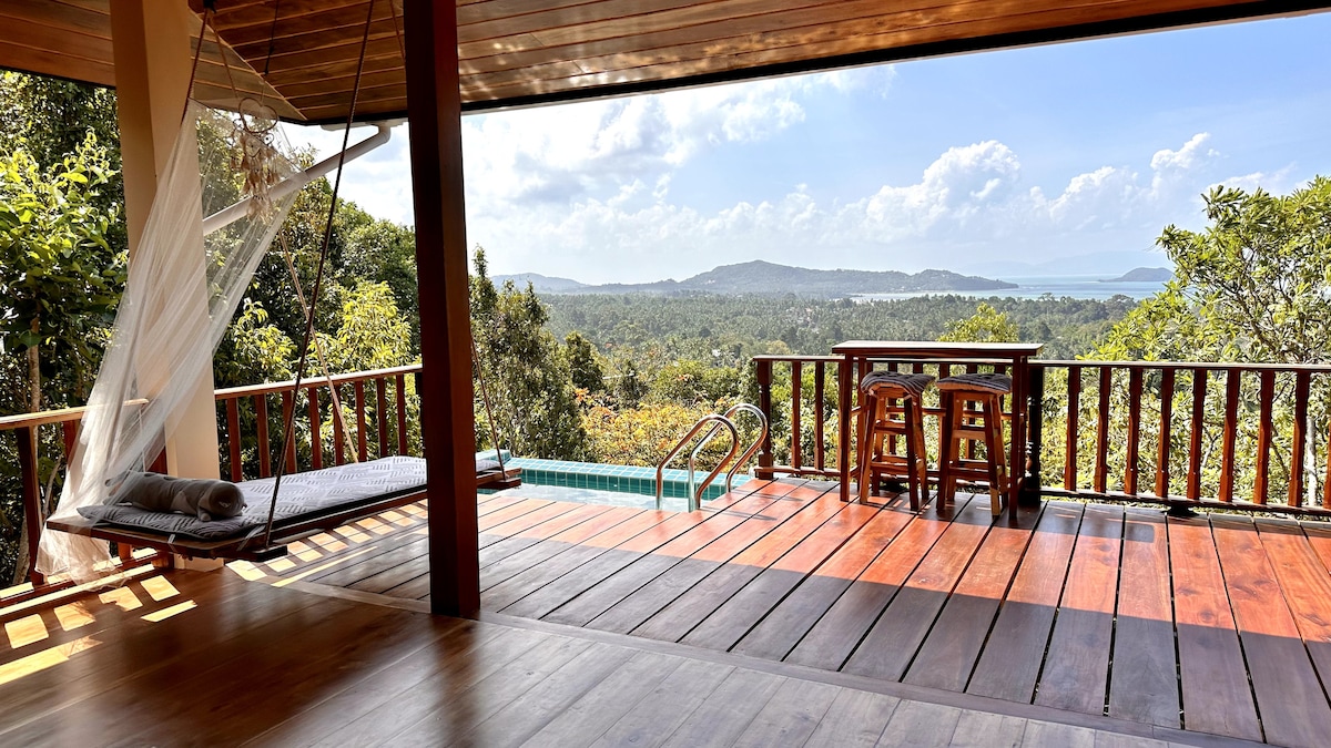 A swinging bed is positioned on the spacious terrace, overlooking a serene landscape featuring lush greenery and distant hills. A swimming pool is visible, alongside a wooden dining area with two chairs and a table, providing a tranquil setting for relaxation.