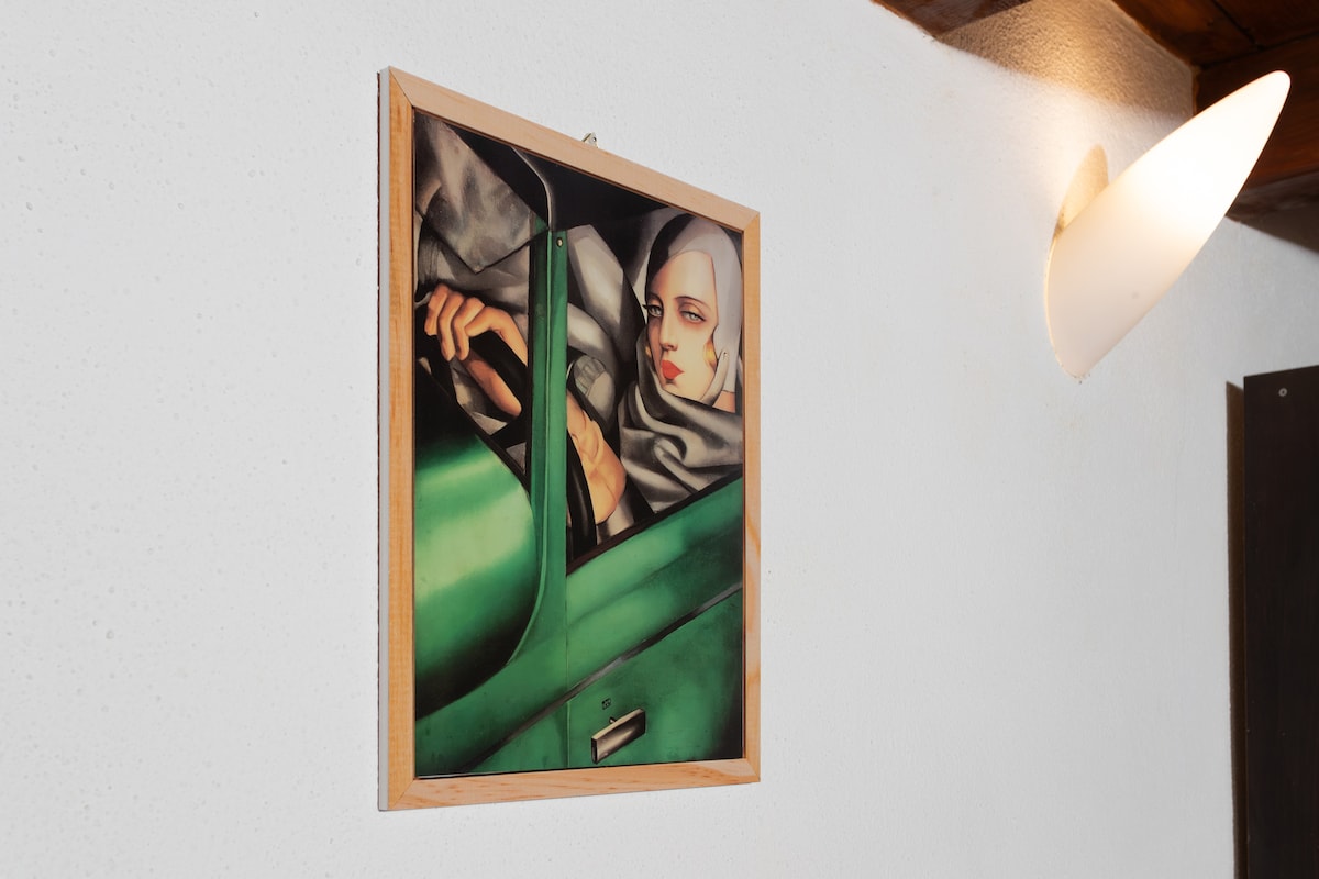 A framed artwork is displayed on the wall, depicting a close-up view of a hand resting on a green vehicle door. Soft lighting from the nearby wall sconce enhances the overall ambiance.