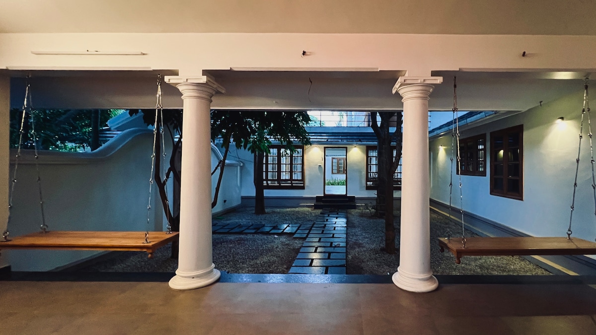 Two wooden swings are suspended from white columns, framing an outdoor courtyard. The courtyard features smooth stone paths, lush greenery, and wooden framed windows illuminating the serene space. A peaceful ambiance is created, encouraging relaxation in the inviting surroundings.