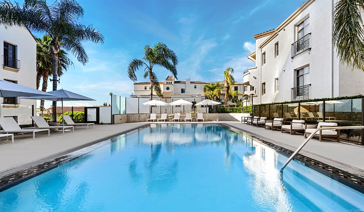 A large swimming pool is surrounded by lounge chairs and shaded by white umbrellas. Lush palm trees provide a tropical feel, and modern buildings with balconies are visible in the background. Clear blue skies enhance the serene setting.