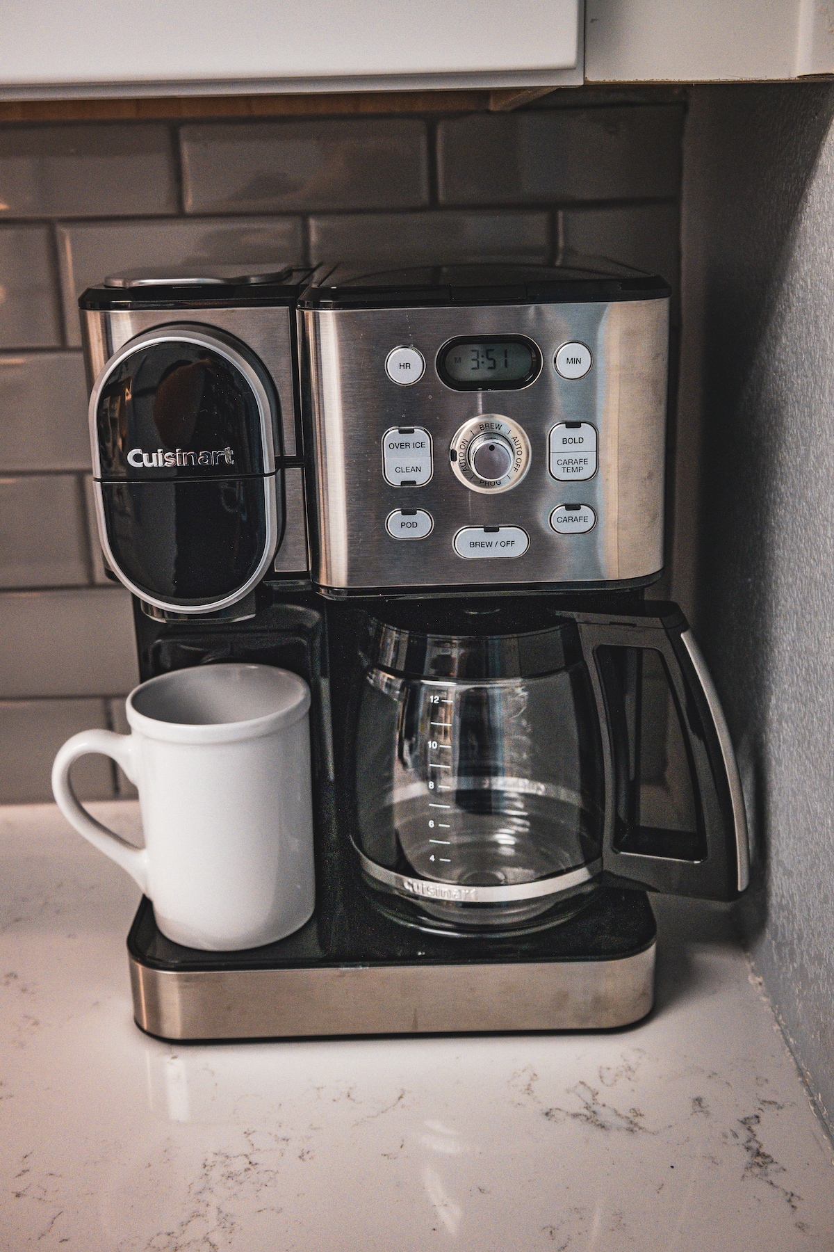 A modern coffee maker is positioned on a countertop, featuring a drip brew system alongside a single-serve option. A white coffee mug is placed next to the glass carafe, ready for use. The surrounding backsplash is made of gray tiles.