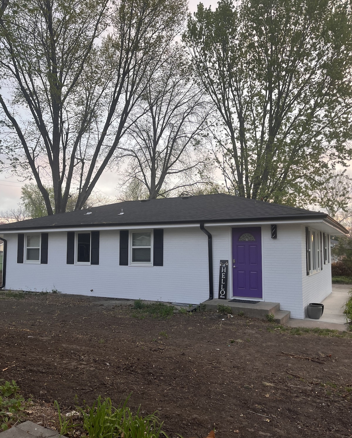 The exterior of the remodeled home features a fresh white brick façade and a dark shingled roof. A welcoming purple front door is framed by black shutters on either side, with trees providing a natural backdrop.