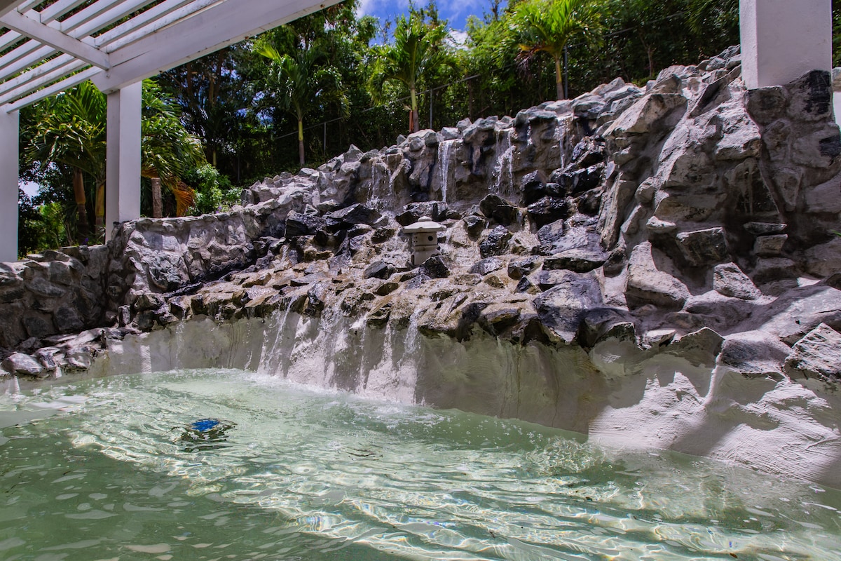 A tranquil pool area is surrounded by natural stone formations, featuring a gentle waterfall that cascades into the clear water. Lush greenery is visible in the background, enhancing the serene atmosphere of the space.