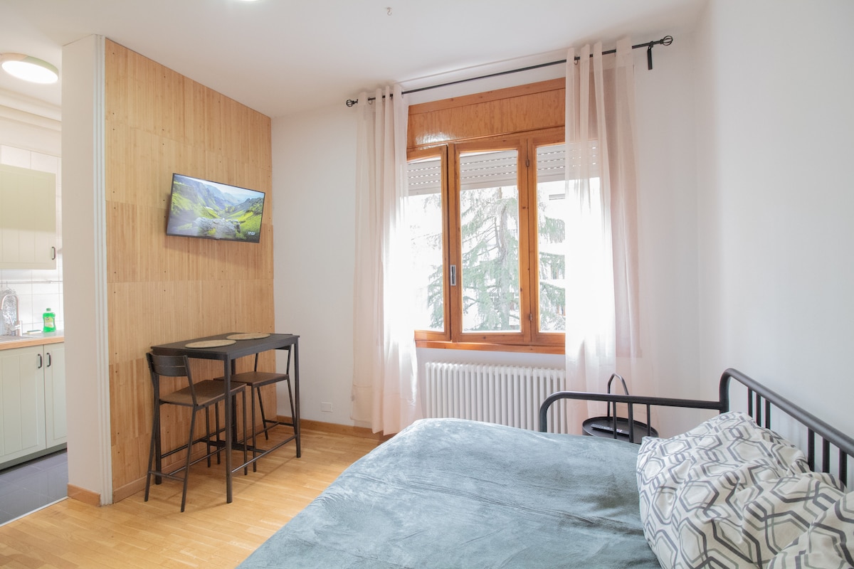 A bright living area is depicted, featuring a spacious bed and a small table with two chairs. A wall-mounted TV displays a scenic landscape, while light filters through the large windows adorned with light curtains, enhancing the warm ambiance of the room.