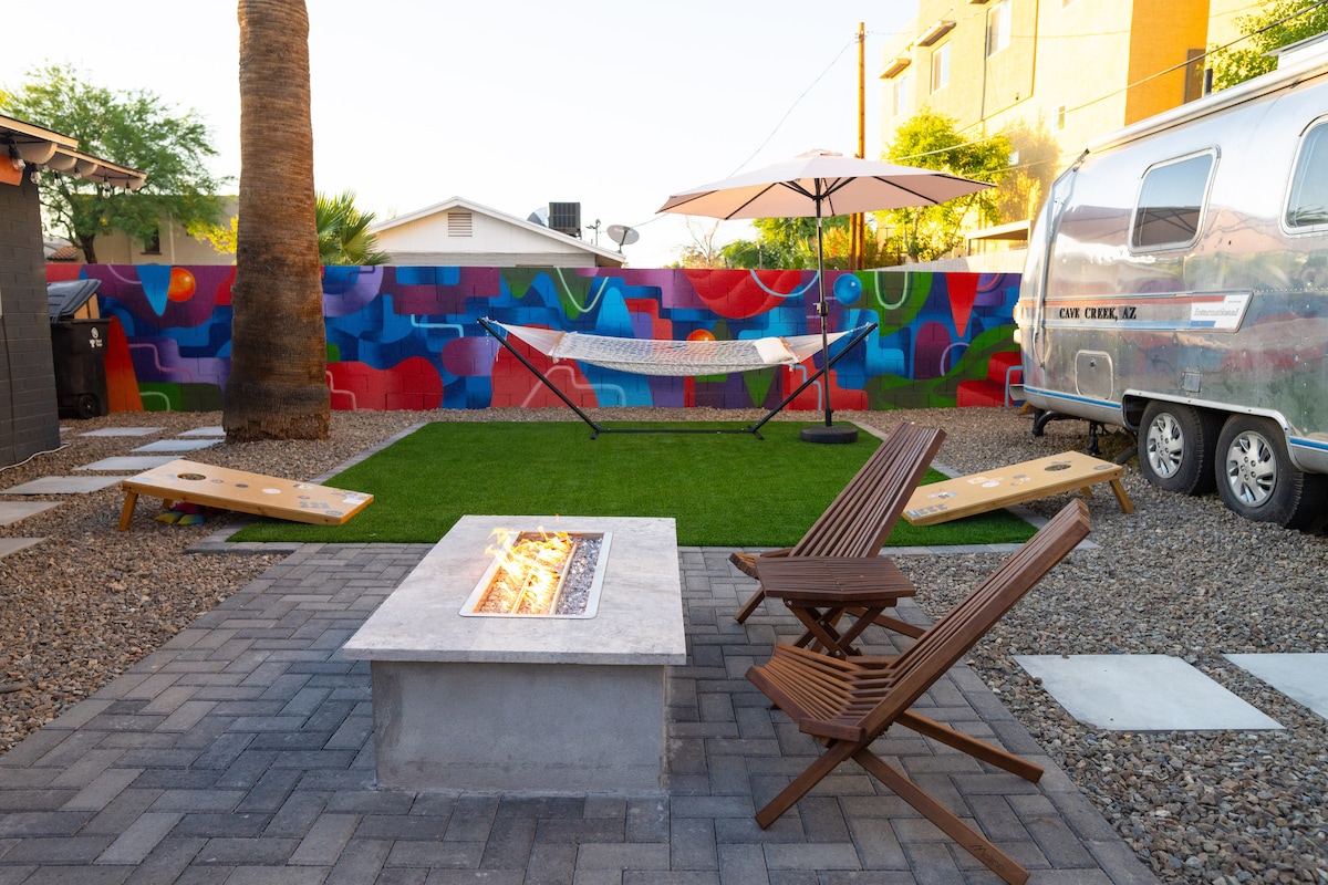 A vibrant outdoor space features a colorful mural as a backdrop. A gas fire pit is surrounded by two wooden chairs on a paver patio. A hammock and an umbrella shade provide relaxation options, while a vintage Airstream adds character to the setting.