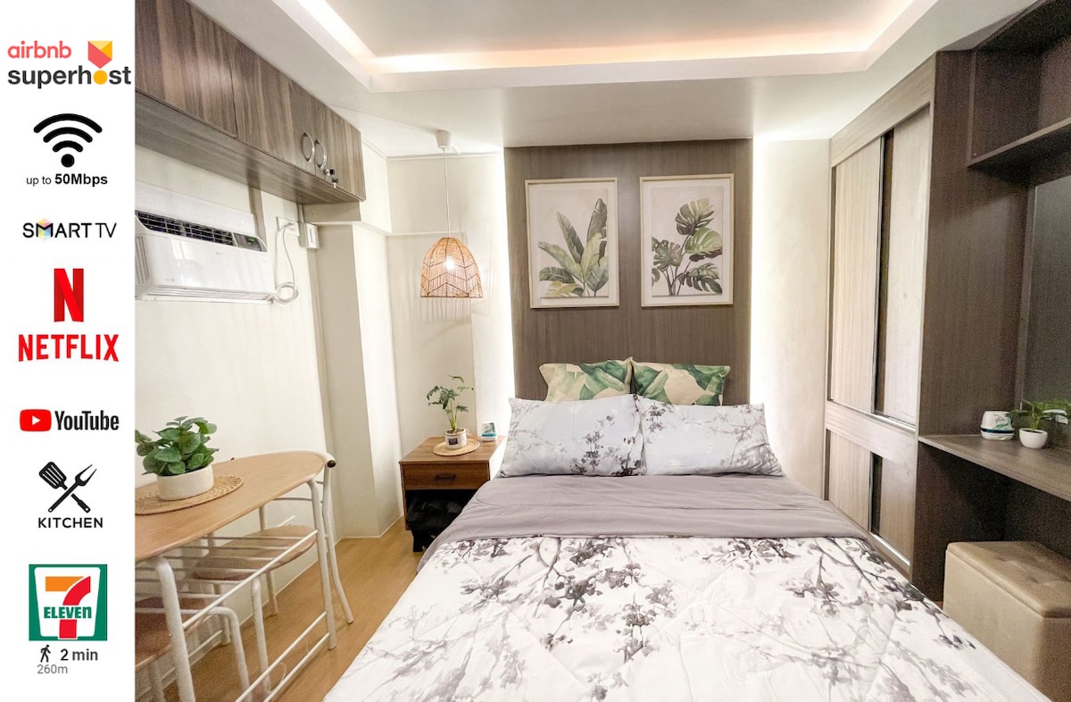A cozy bedroom area features a double size bed dressed in soft linens, surrounded by nuanced wall colors. Contemporary art pieces hang above the bed. A small table and chairs are situated nearby, providing a workspace, while warm lighting enhances the inviting atmosphere.