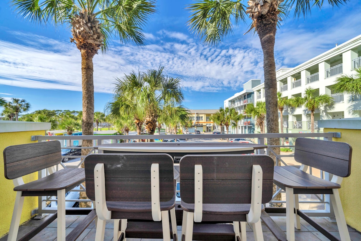 An oversized private balcony is shown, equipped with four chairs and surrounded by palm trees. The vibrant blue sky and wispy clouds create a clear backdrop, while shops and artist booths are visible below.