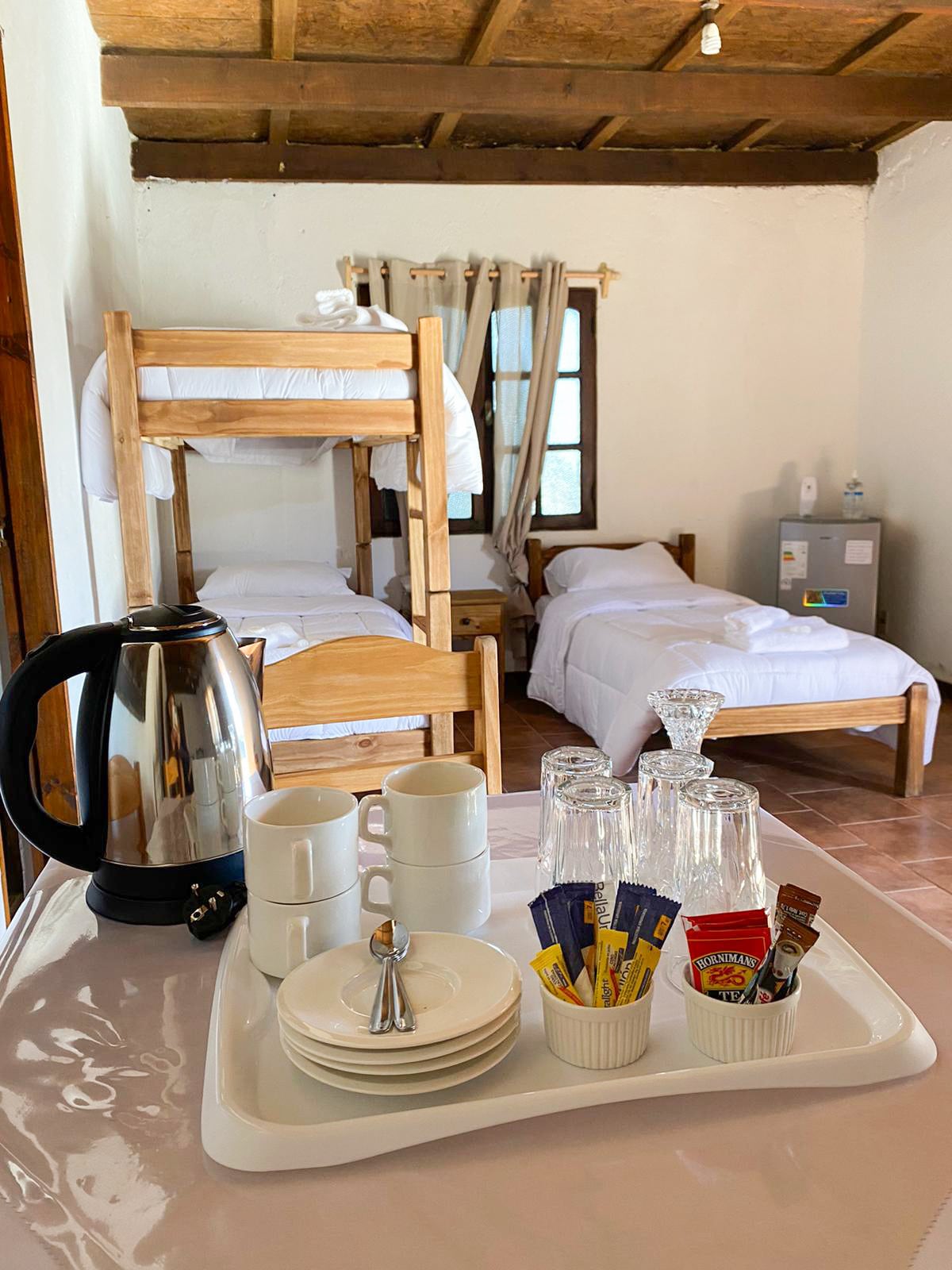 A cozy sleeping area is visible, featuring a bunk bed and a single bed. A table is set with a kettle, cups, and glasses, along with a selection of tea and sugar packets. Natural light enters through a window with light curtains.