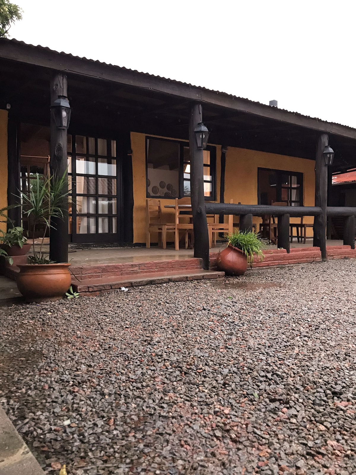 The exterior of the building is characterized by warm, earthy tones and rustic wooden beams. A covered porch provides seating options, framed by potted plants. A gravel walkway leads up to the entrance, where large windows invite natural light into the interior.
