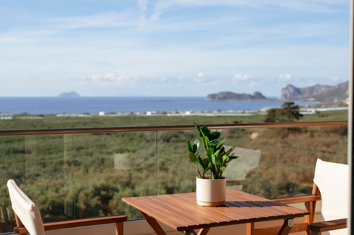 A wooden table is positioned on a balcony, accompanied by two light-colored chairs. A small potted plant sits at the center of the table. The view encompasses the sea and distant landforms, with a clear sky above, creating a serene outdoor setting.