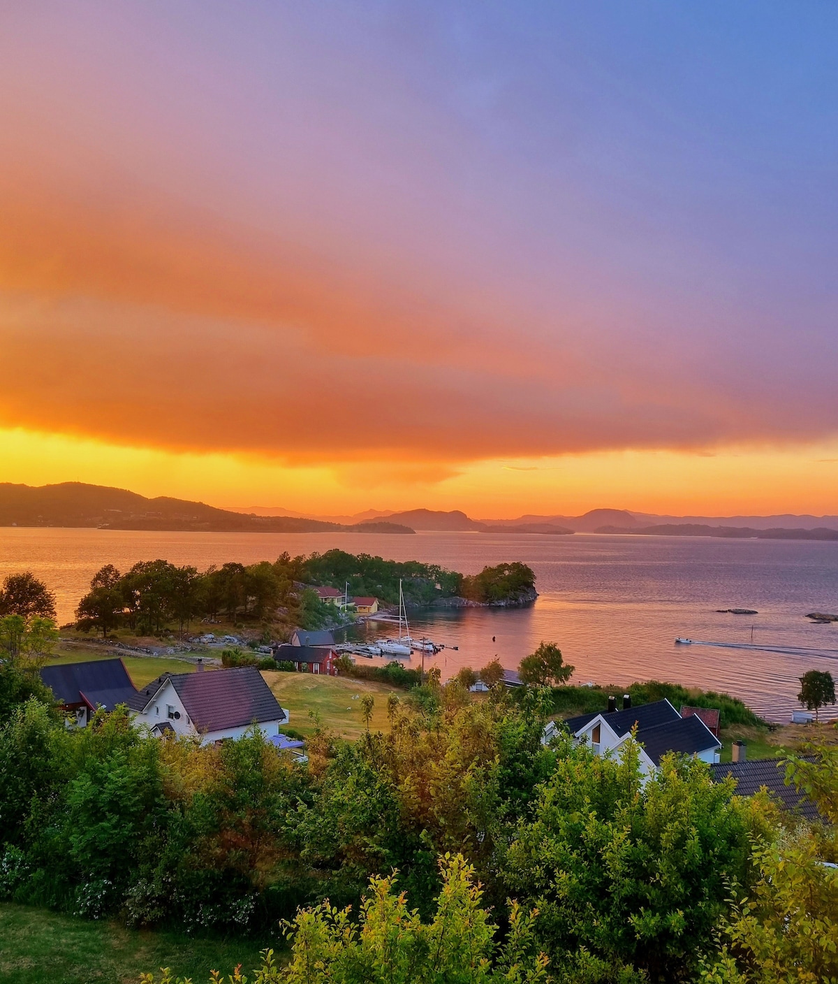 A scenic view of a tranquil waterfront at sunset, featuring a range of houses along the shoreline. The sky is illuminated with warm hues of orange and purple. Lush green vegetation and distant hills are also visible, enhancing the natural beauty of the surrounding landscape.