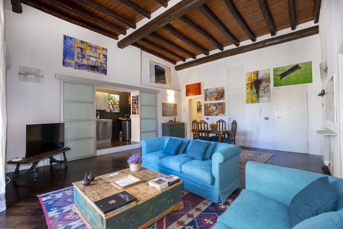 A spacious living area features high wooden beams overhead, creating an airy ambiance. Comfortable turquoise sofas are arranged around a rustic coffee table. Artworks adorn the walls, adding visual interest. The dining area and kitchen are visible through a glass door, contributing to an open and inviting layout.