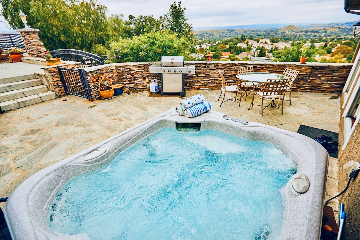 A hot tub is situated on a stone patio surrounded by an inviting landscape and scenic views. Nearby, a BBQ grill and a circular table with chairs offer comfortable outdoor dining options. The peaceful atmosphere is enhanced by surrounding greenery.