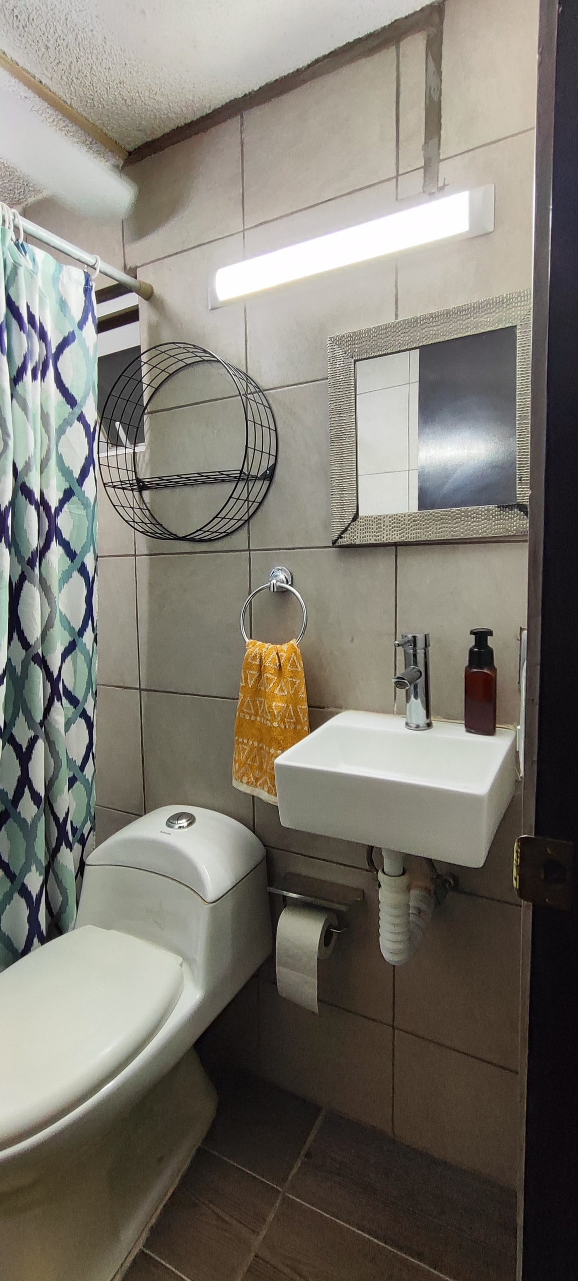 A compact bathroom features light-colored tiles and a modern sink. A decorative mirror and a wall-mounted basket add to the sleek design. A bright colored towel hangs beside the sink, offering a pop of color, while a curtain provides privacy.