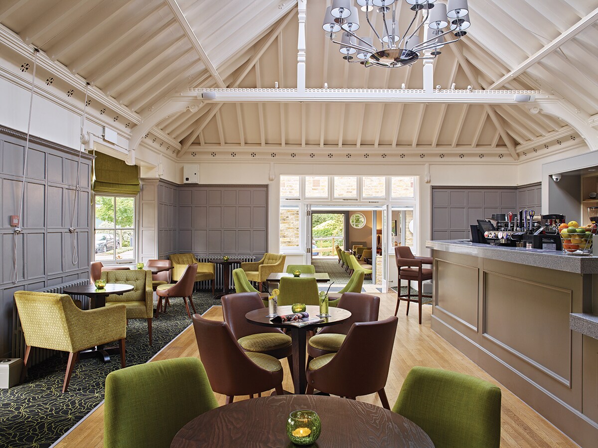 An inviting lounge area features high ceilings with wooden beams and a stylish chandelier. Tables are arranged with green and brown upholstered chairs, creating a relaxed atmosphere. Natural light pours in through large windows, illuminating the space with a warm glow.
