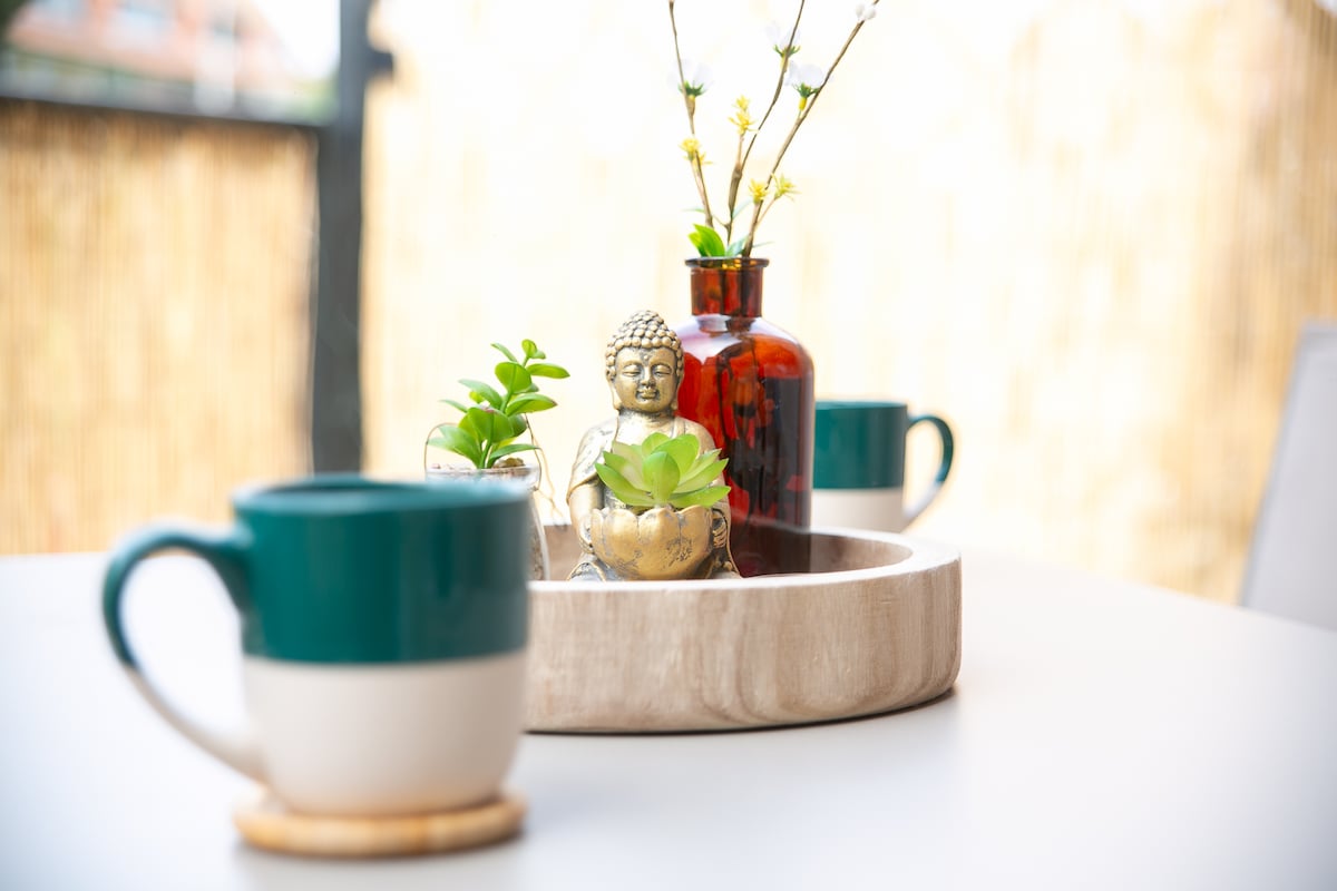 A round wooden tray is decorated with small plants and a decorative bottle, creating a serene tabletop display. Two coffee mugs are positioned on either side, complementing the arrangement. The background features a natural, textured screen, enhancing the outdoor ambiance.