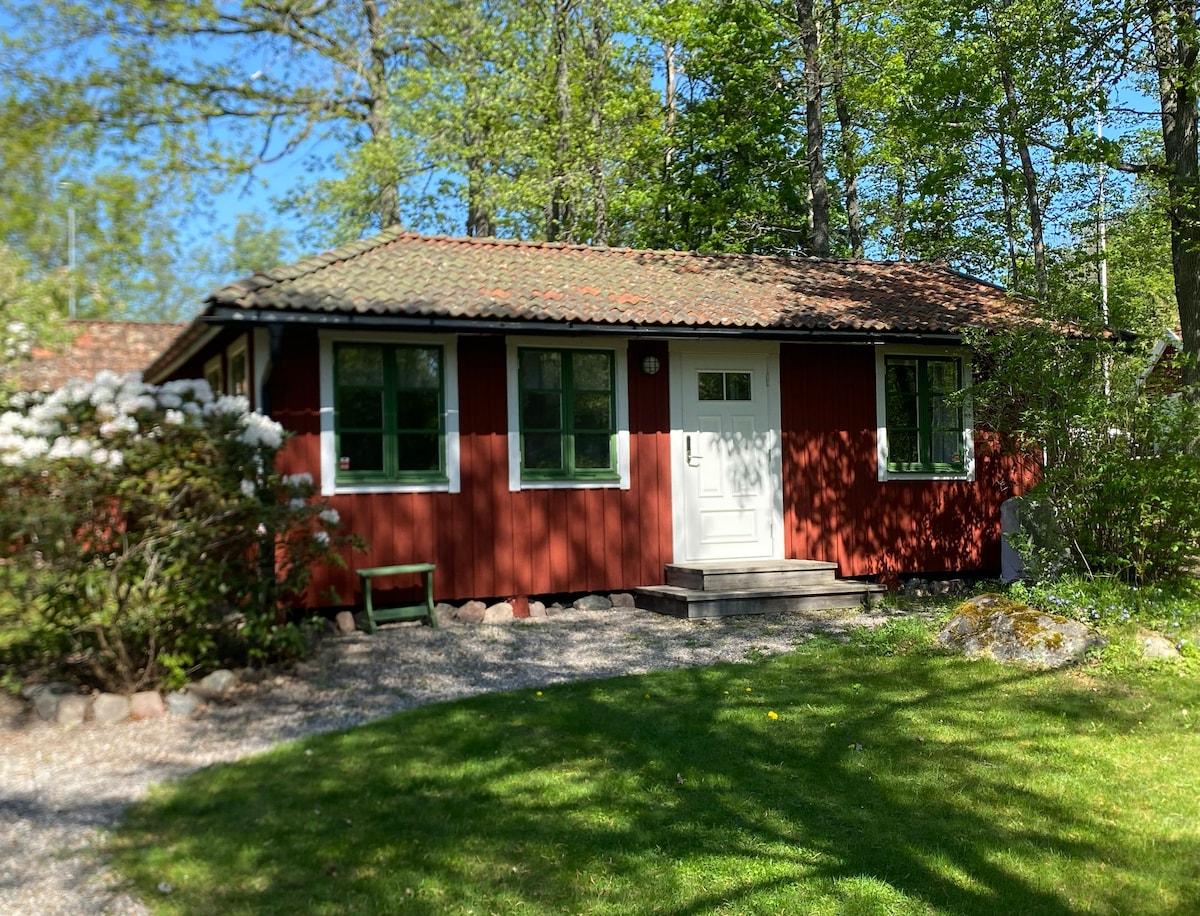 A charming red cottage is set within a green landscape, surrounded by trees and colorful flowering bushes. The front door is centered and framed by two windows, creating a welcoming entrance. A stone pathway leads to the entrance, enhancing the natural surroundings.
