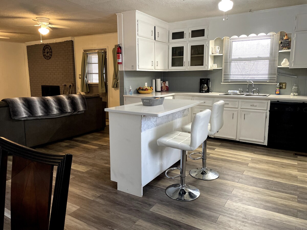 A spacious kitchen features a central island with seating for three. White cabinetry lines the walls, complemented by a gray backsplash. A coffee maker and various kitchen utensils are visible. The open layout extends into the living area, where a comfortable sofa is positioned.