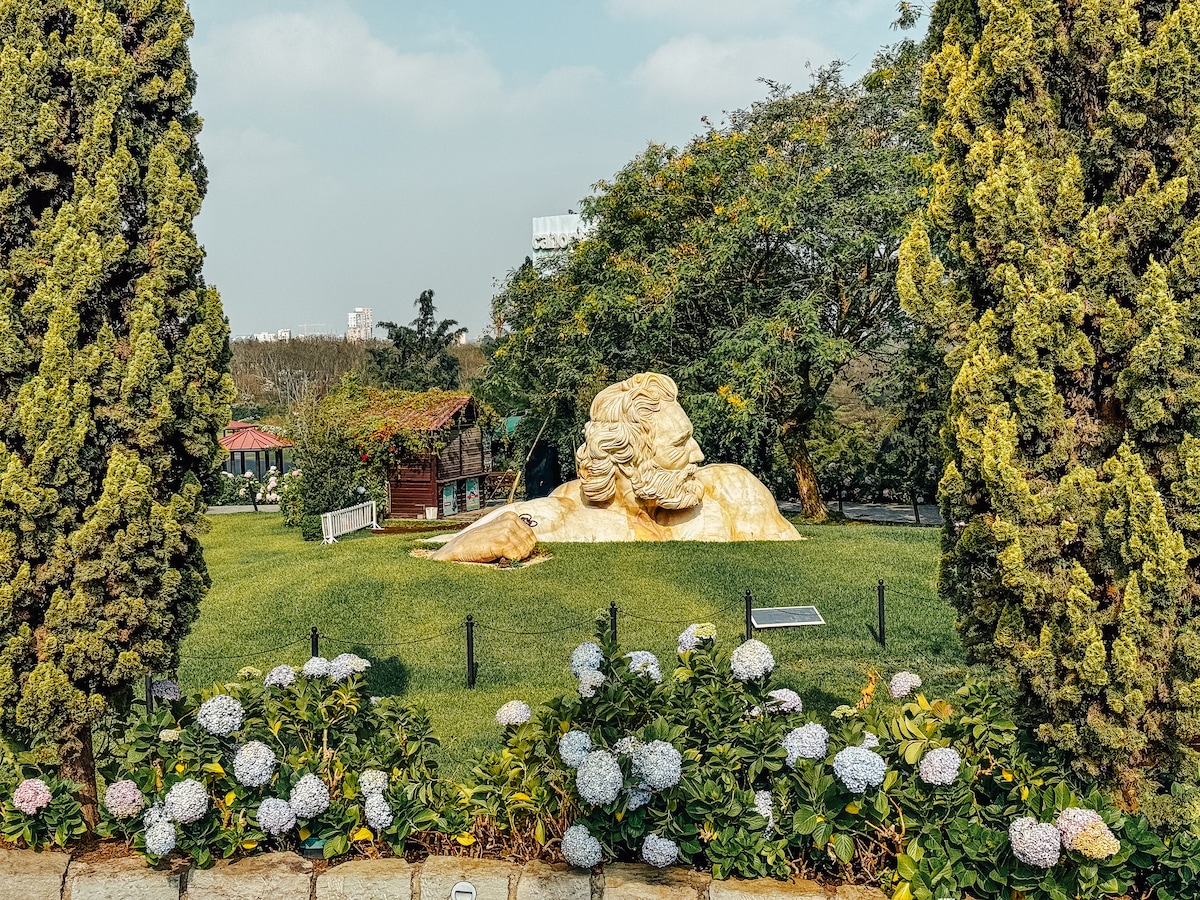 A large golden sculpture of a reclining figure is positioned in a lush green garden, surrounded by vibrant flowers and manicured shrubs. Towers of greenery rise on both sides, framing the sculpture and enhancing the serene outdoor atmosphere.