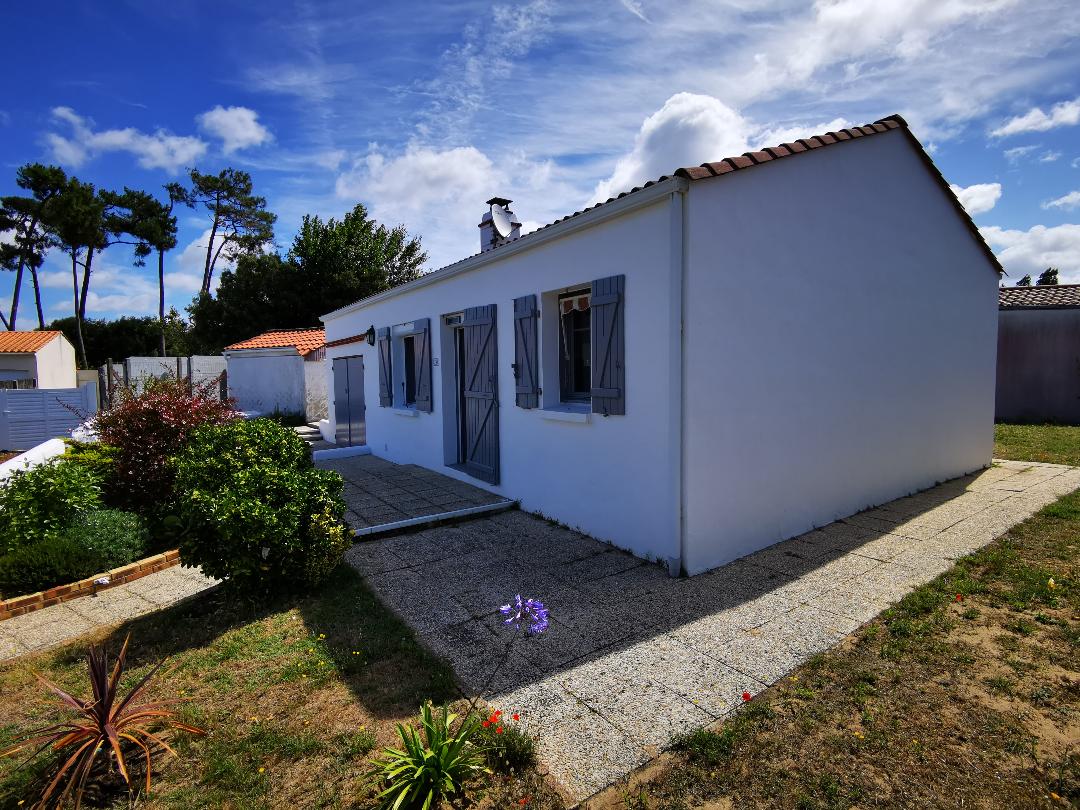 The exterior of a single-story white house is depicted, featuring grey shutters and a simple front entrance. A pathway made of gravel leads to the house, with patches of grass and scattered flowers enhancing the outdoor space. Trees are visible in the background under a partly cloudy sky.