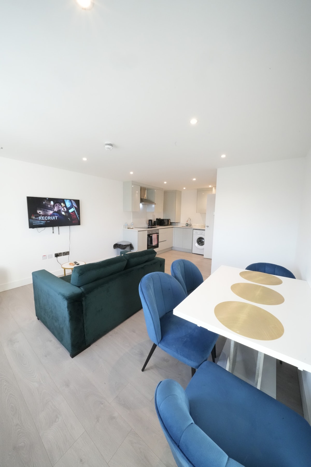 A modern living area features a green velvet sofa facing a wall-mounted television. A dining table with gold accents and blue chairs is positioned nearby. The open kitchen is visible in the background, equipped with stainless steel appliances and cabinets.