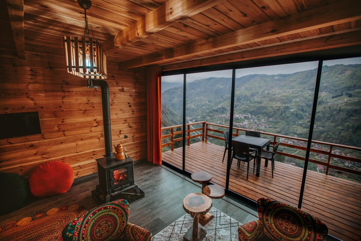A warm living area features wooden walls and ceiling, with a cozy stove providing warmth. Large glass doors open to a spacious balcony with views of the surrounding mountains. Rattan chairs and a patterned table create a comfortable sitting area, inviting relaxation.