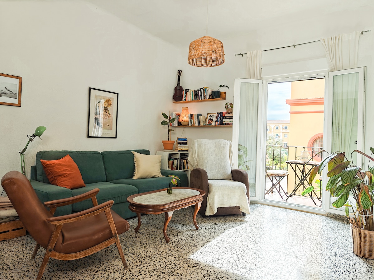 A welcoming living area features a comfortable green sofa paired with two armchairs. Natural light fills the space through open double doors leading to a small balcony with a table and chairs. A wooden coffee table rests at the center, surrounded by indoor plants and bookshelves.