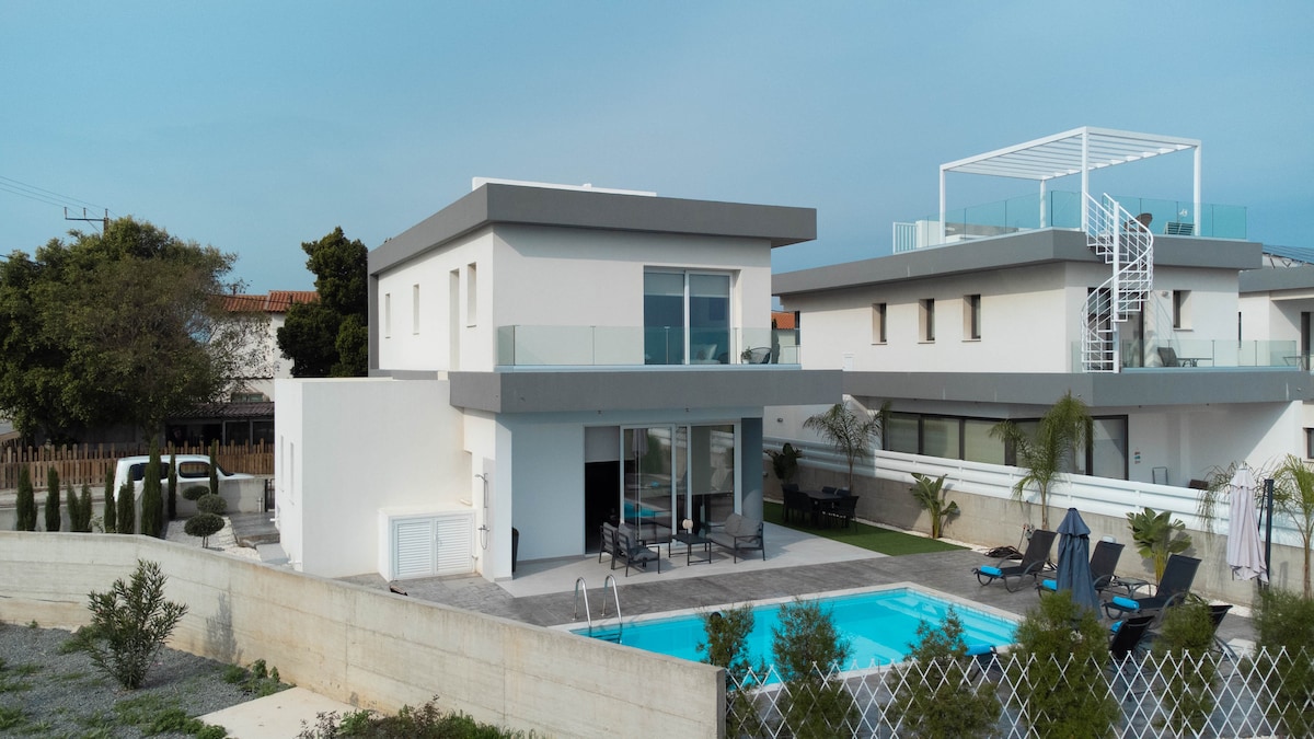 A modern two-story villa is presented with a sleek design, featuring large windows and a private swimming pool surrounded by lounge chairs. The outdoor area is landscaped, providing a welcoming atmosphere, while a spacious terrace offers additional seating and relaxation options.
