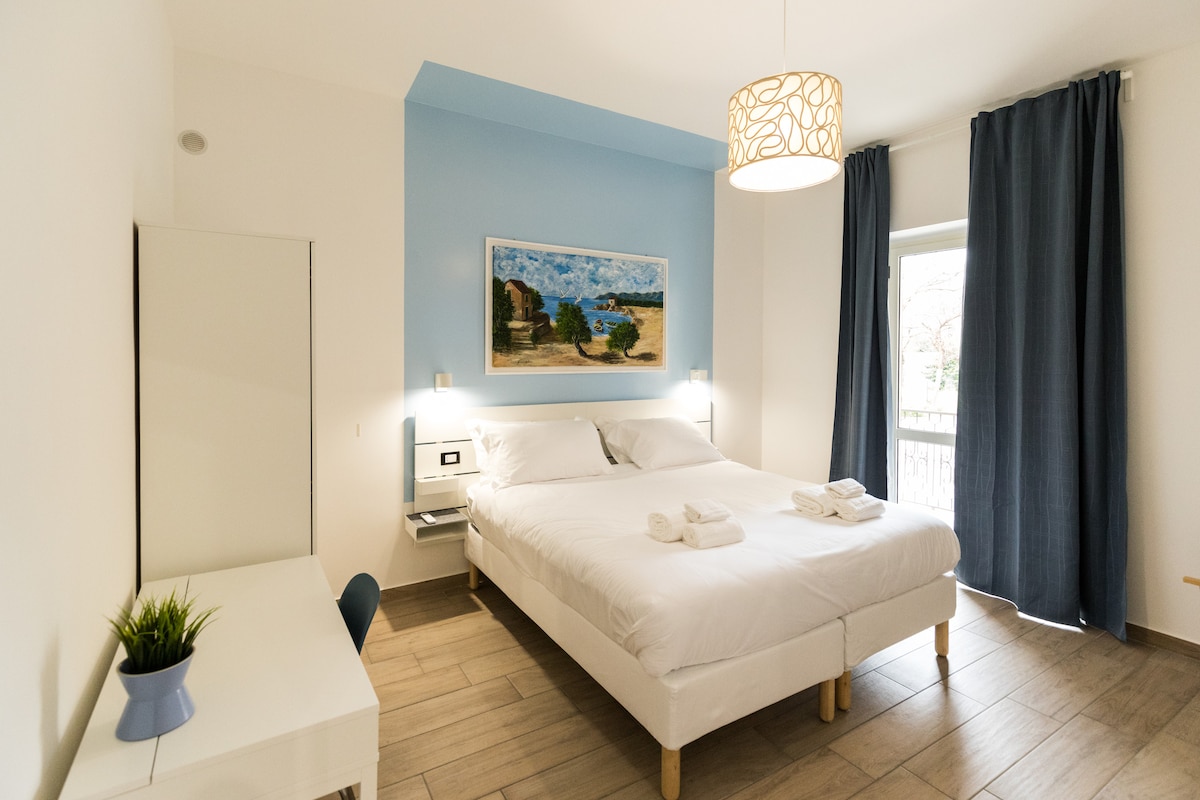 A bright room features a spacious bed with crisp white linens, complemented by a light blue accent wall. A painting hangs above the bed, and a desk sits nearby. Large curtains frame the window, allowing natural light to fill the space.