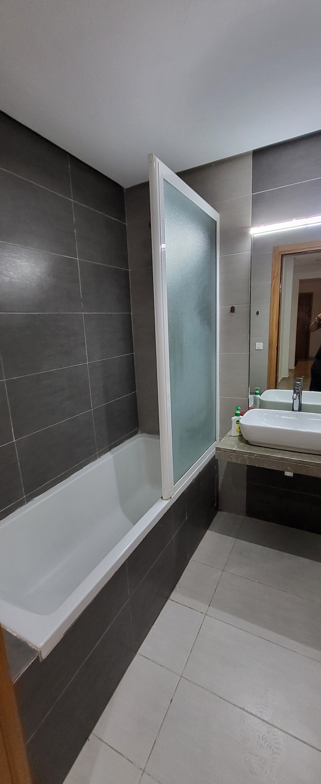 The bathroom features a clean, modern design with neutral grey tiles and a glass shower screen. A spacious bathtub is situated alongside a vanity with a sleek countertop and a mirror, providing a functional and tidy space.
