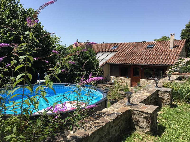 A swimming pool can be seen surrounded by lush greenery and colorful flowers, while a stone wall frames the area. A rustic building with a red tiled roof is visible in the background, featuring large windows and a terrace for outdoor seating.