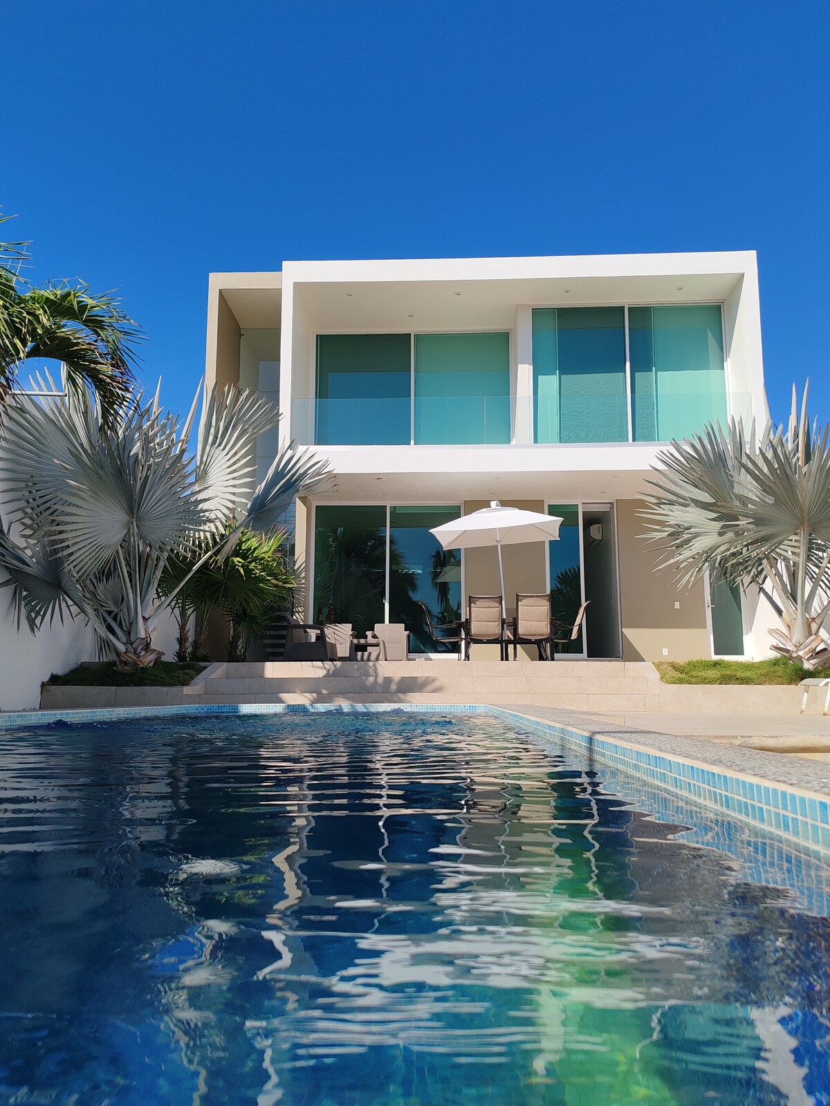 A modern two-story house features large glass windows that reflect the clear blue sky. An inviting swimming pool is visible in the foreground, with a sunshade and outdoor seating area placed conveniently on the terrace, surrounded by lush palm foliage.