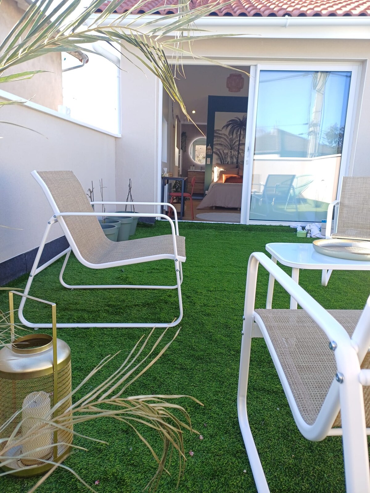 A bright terrace features artificial grass, creating a green and inviting atmosphere. Two white chairs and a small table are positioned on the patio, alongside a lantern and potted plants. Sunlight filters in through the glass doors, illuminating the space.