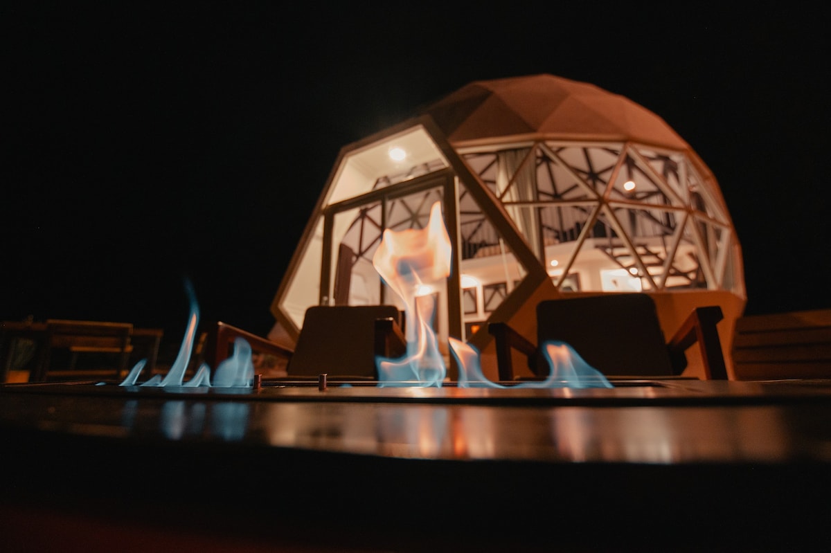 A geodesic dome is illuminated at night, showcasing its unique architectural design with large glass windows. In the foreground, a fire pit displays lively flames, providing a warm glow against the dark backdrop, with comfortable seating arranged nearby.